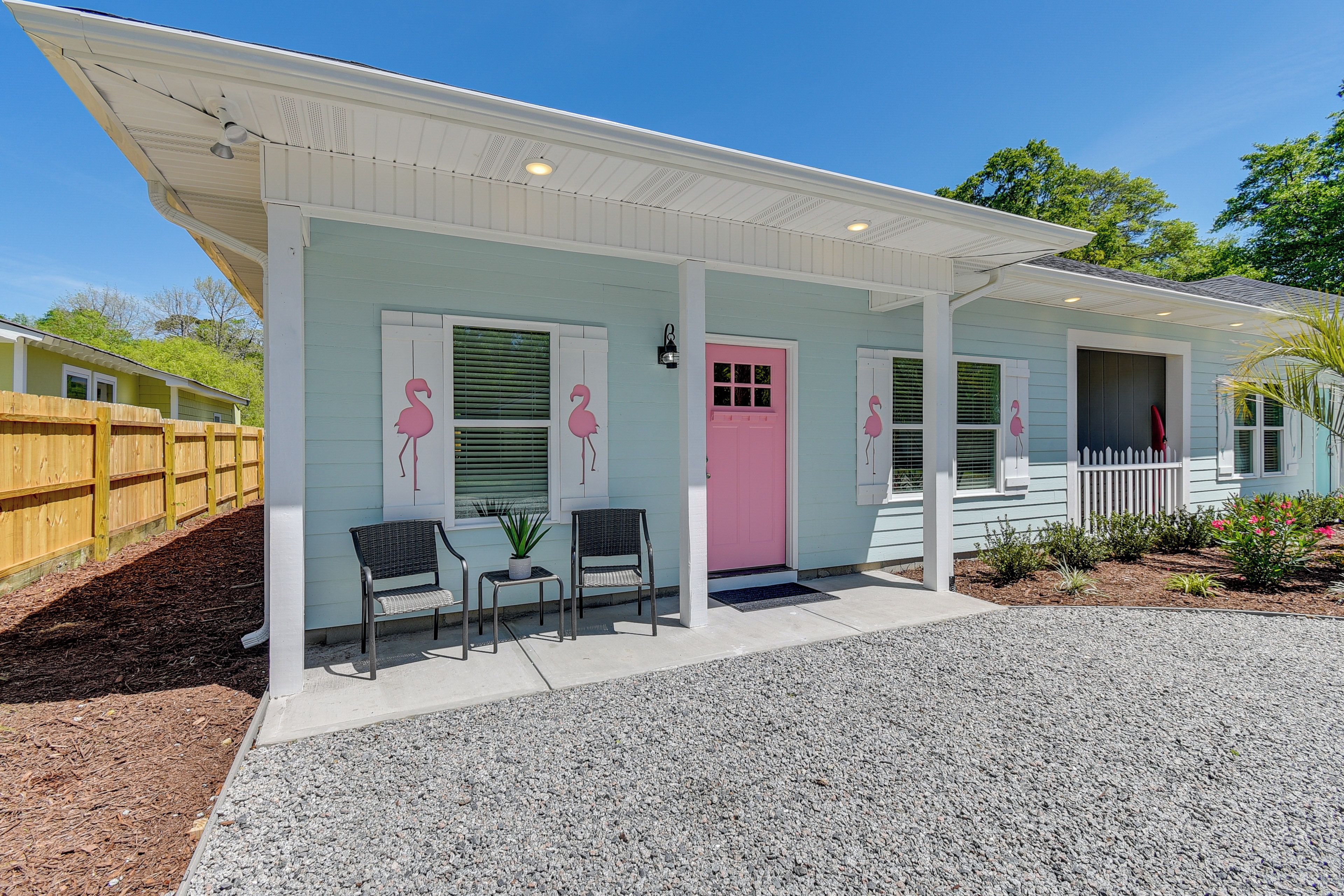 A light blue house with a pink door and windows, a covered porch with two chairs and a table, and a wooden fence in the background.