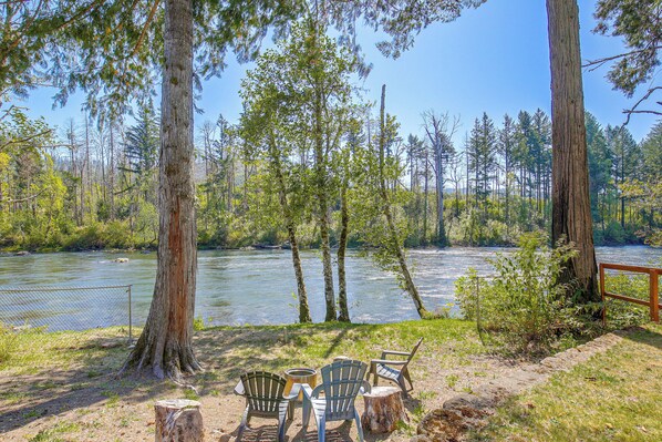 A riverside campsite with chairs and a table, surrounded by trees and a wooden fence.