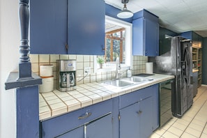 A kitchen with blue cabinets, a coffee maker, a refrigerator, and a window with a view of trees.