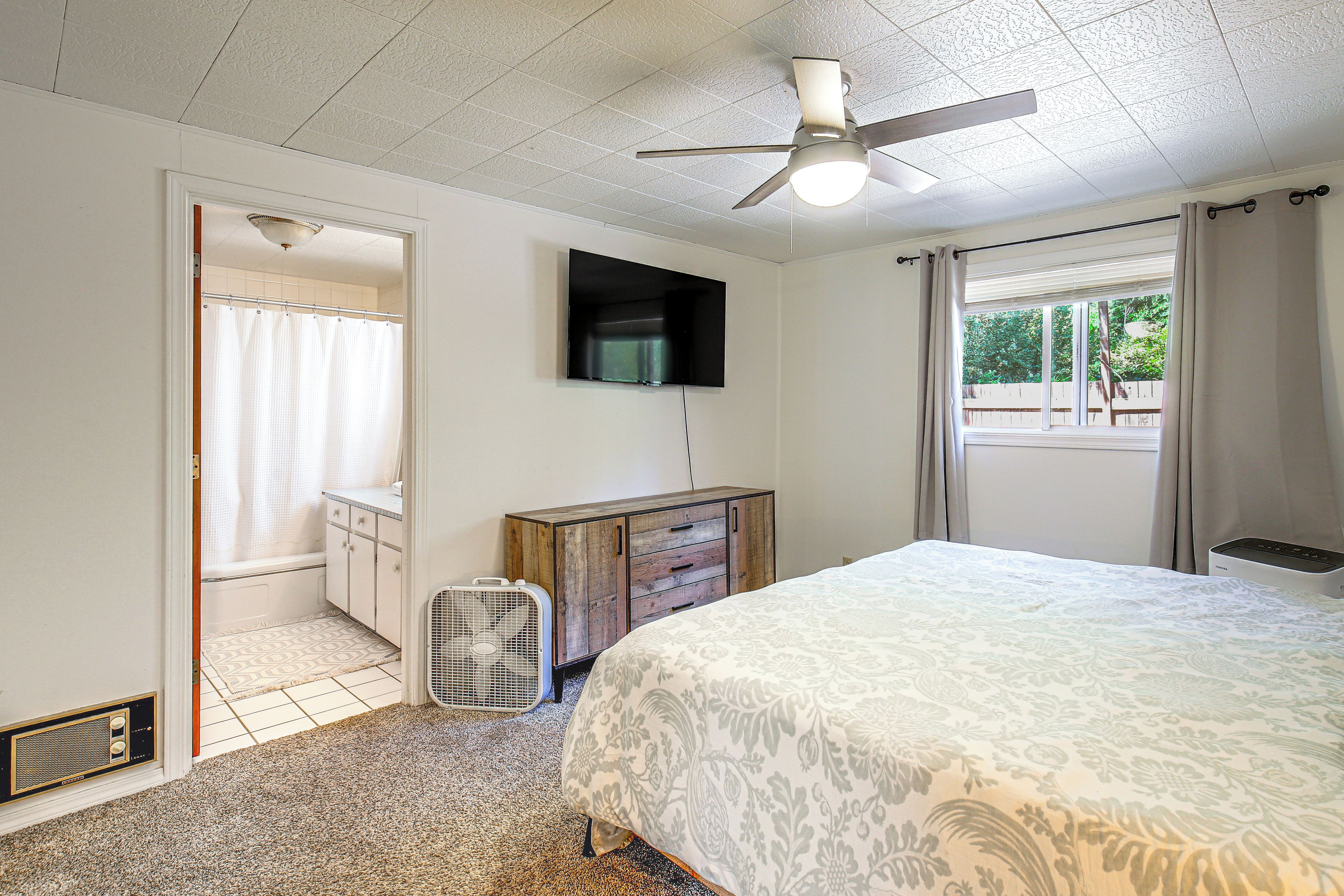 Ein Schlafzimmer mit einem Bett, einem Deckenventilator, einem Fernseher, einer Heizung und einem Fenster mit Vorhängen.