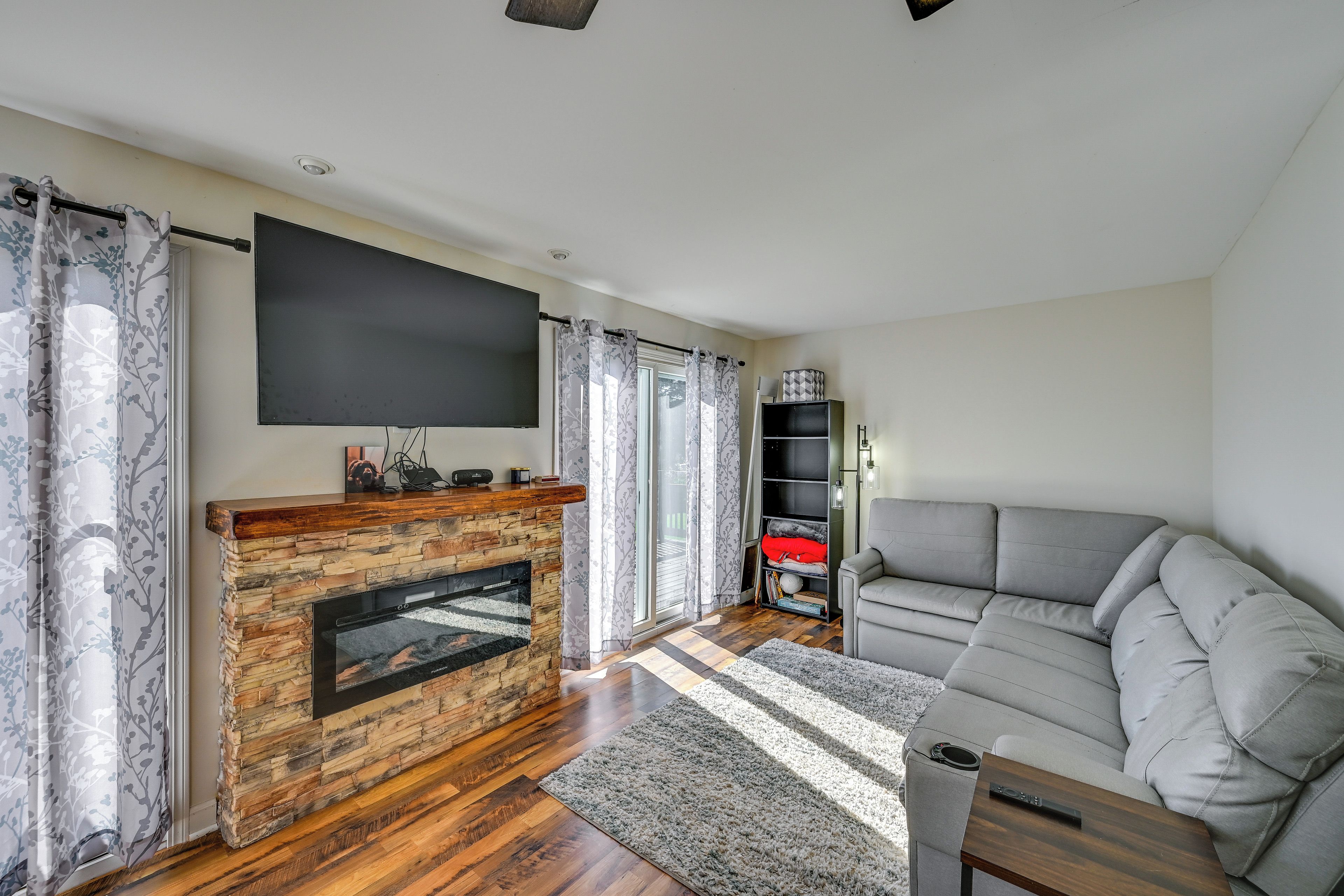 A modern living room with a stone fireplace, a flat-screen TV, grey sofa, and a bookshelf.