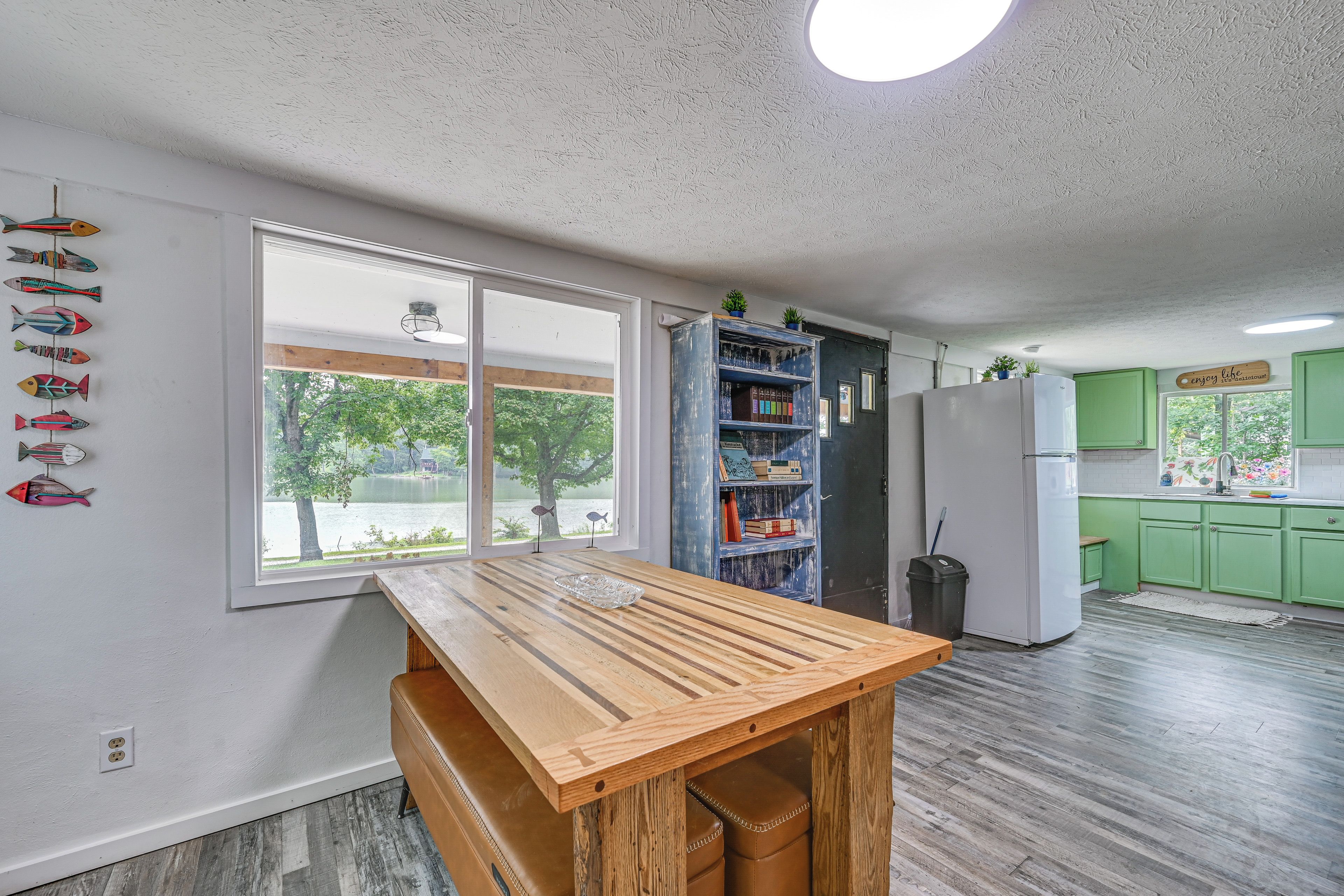 Una cocina con mesa de madera, armarios verdes y vista a un lago a través de amplios ventanales.