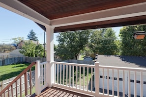 Ein überdachter Balkon mit Holzgeländer, Blick auf einen Garten mit Zaun und ein Haus im Hintergrund.