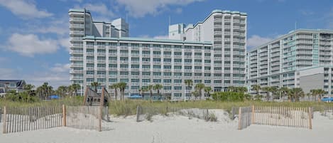 A beachfront hotel with multiple balconies, a sandy beach in the foreground, and a clear sky.