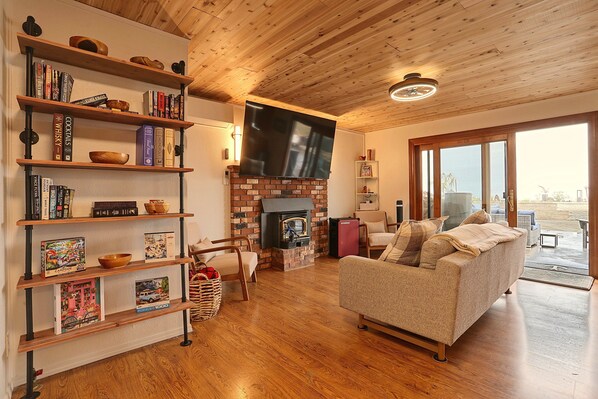 A cozy living room with a fireplace, wooden floors, a flat-screen TV, and a bookshelf filled with books. - Waterfront Bellingham Home w/ Fire Pits & Patio (Bellingham)