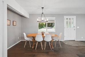 A dining area with a wooden table and white chairs, a chandelier, and a view through a window. - Cabin & Guest House Near Sandy River Trails! (Brightwood)