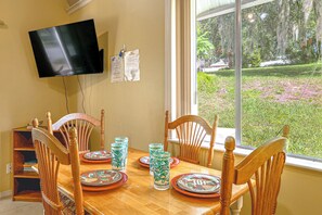A dining area with a wooden table set for four, a television mounted on the wall, and a window with a view of a garden. - 'livin' on Sunshine' Lake Louisa Escape! (Clermont)