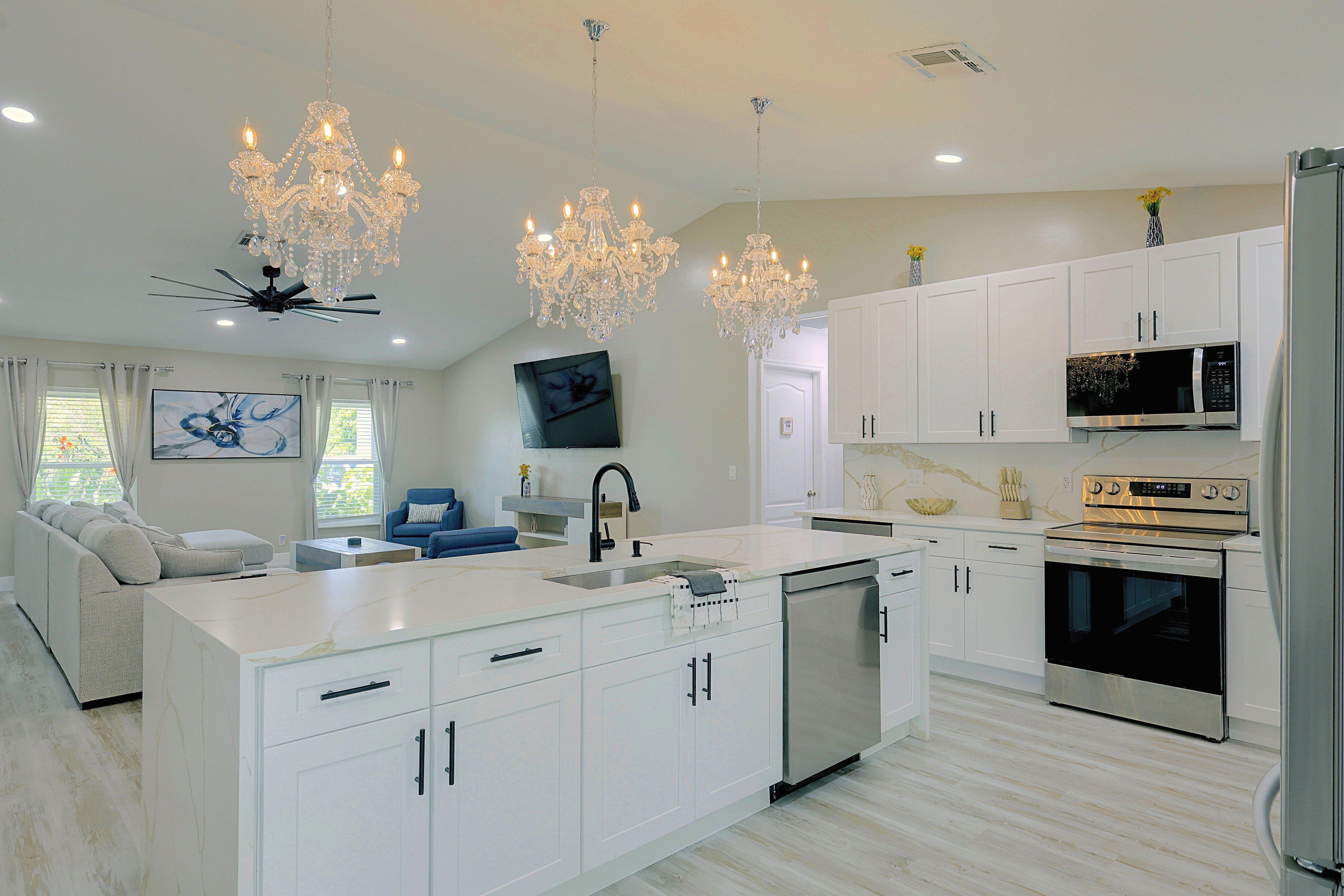 A modern kitchen with white cabinetry, stainless steel appliances, a central island with a sink, and a chandelier.