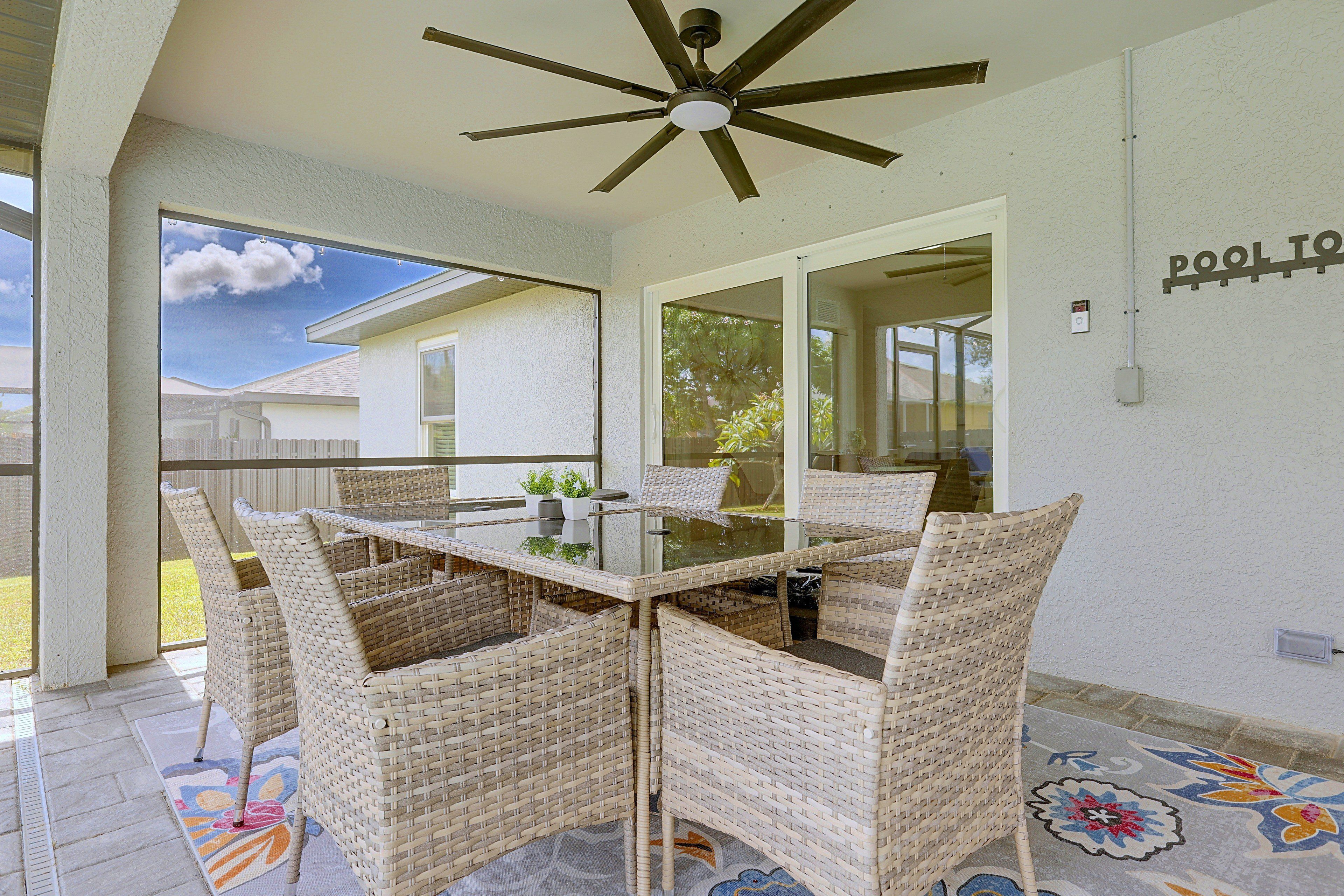 Un patio techado con una mesa de vidrio y sillas de mimbre, un ventilador de techo y vistas a una casa.