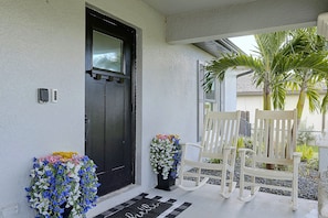 A black door with a window, a white chair, and two flower arrangements on the porch. - Florida Sunshine! Cape Coral Home w/ Pool (Cape Coral)