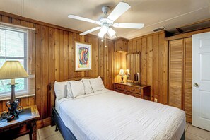 Une chambre à coucher comprenant un lit, un ventilateur de plafond, une lampe de chevet, une commode en bois, un miroir et une fenêtre avec des stores.