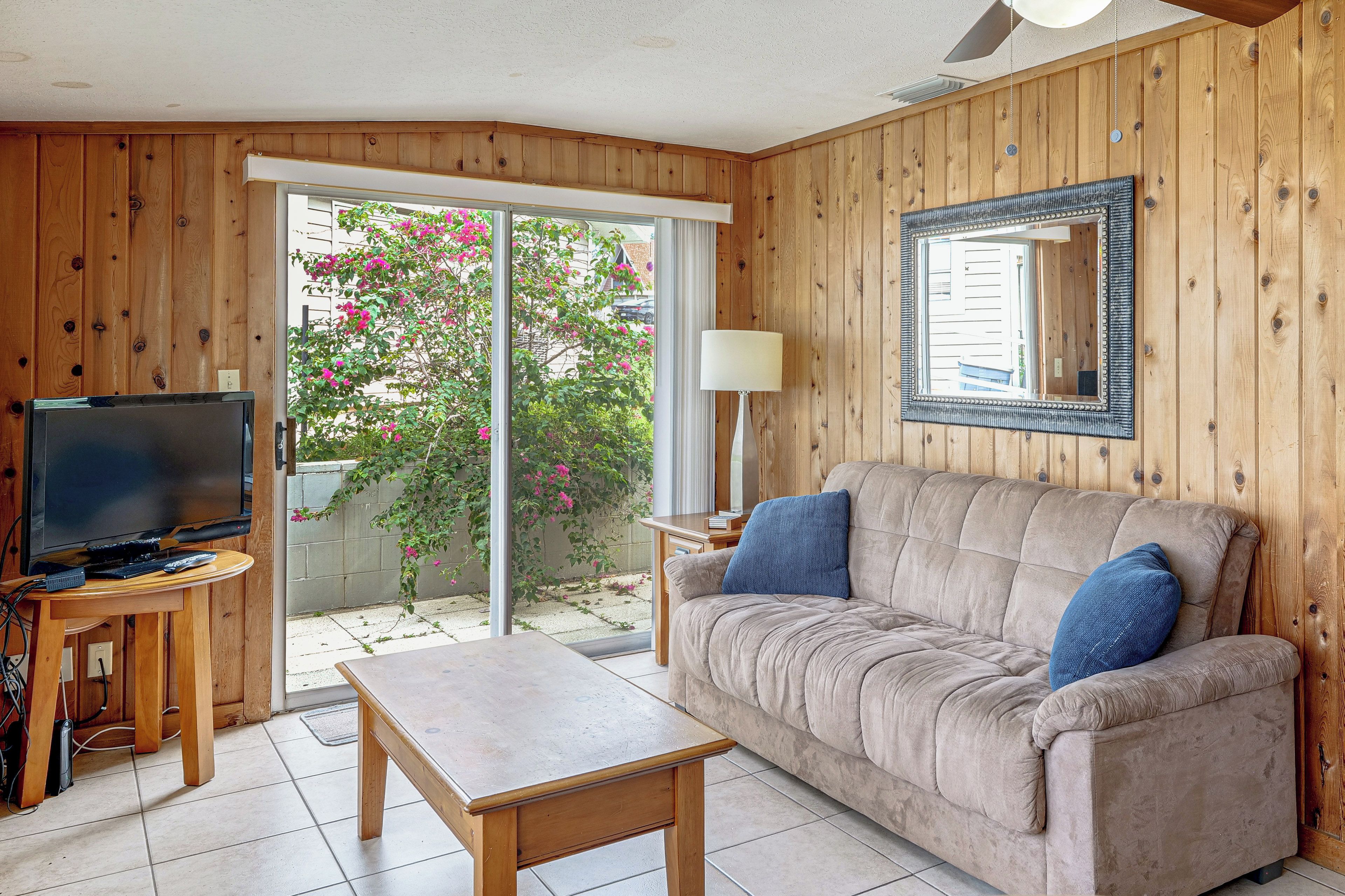 A living room with a television, a sofa, a table, and a sliding glass door leading to a balcony with flowers.