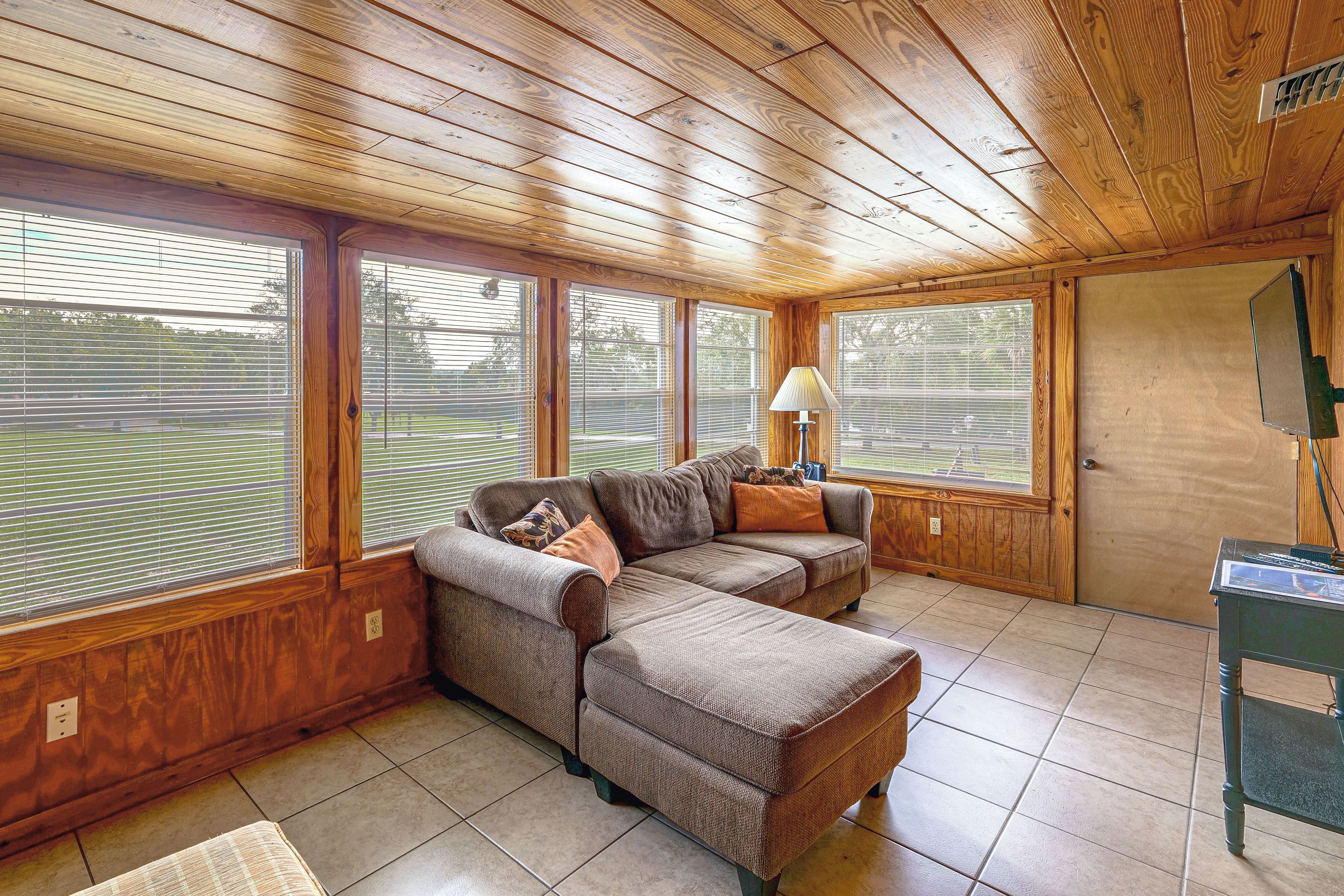 A room with a wooden ceiling, large windows with blinds, a sectional sofa with cushions, and a small table with a lamp.