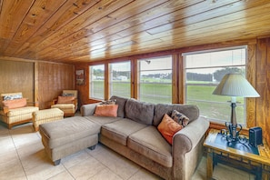 Ein gemütliches Wohnzimmer mit einem Ecksofa, Sesseln und einem kleinen Tisch mit einer Lampe. Große Fenster bieten Blick auf eine grüne Landschaft.