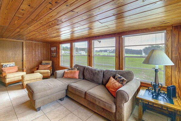 Ein gemütliches Wohnzimmer mit einem Ecksofa, Sesseln und einem kleinen Tisch mit einer Lampe. Große Fenster bieten Blick auf eine grüne Landschaft.