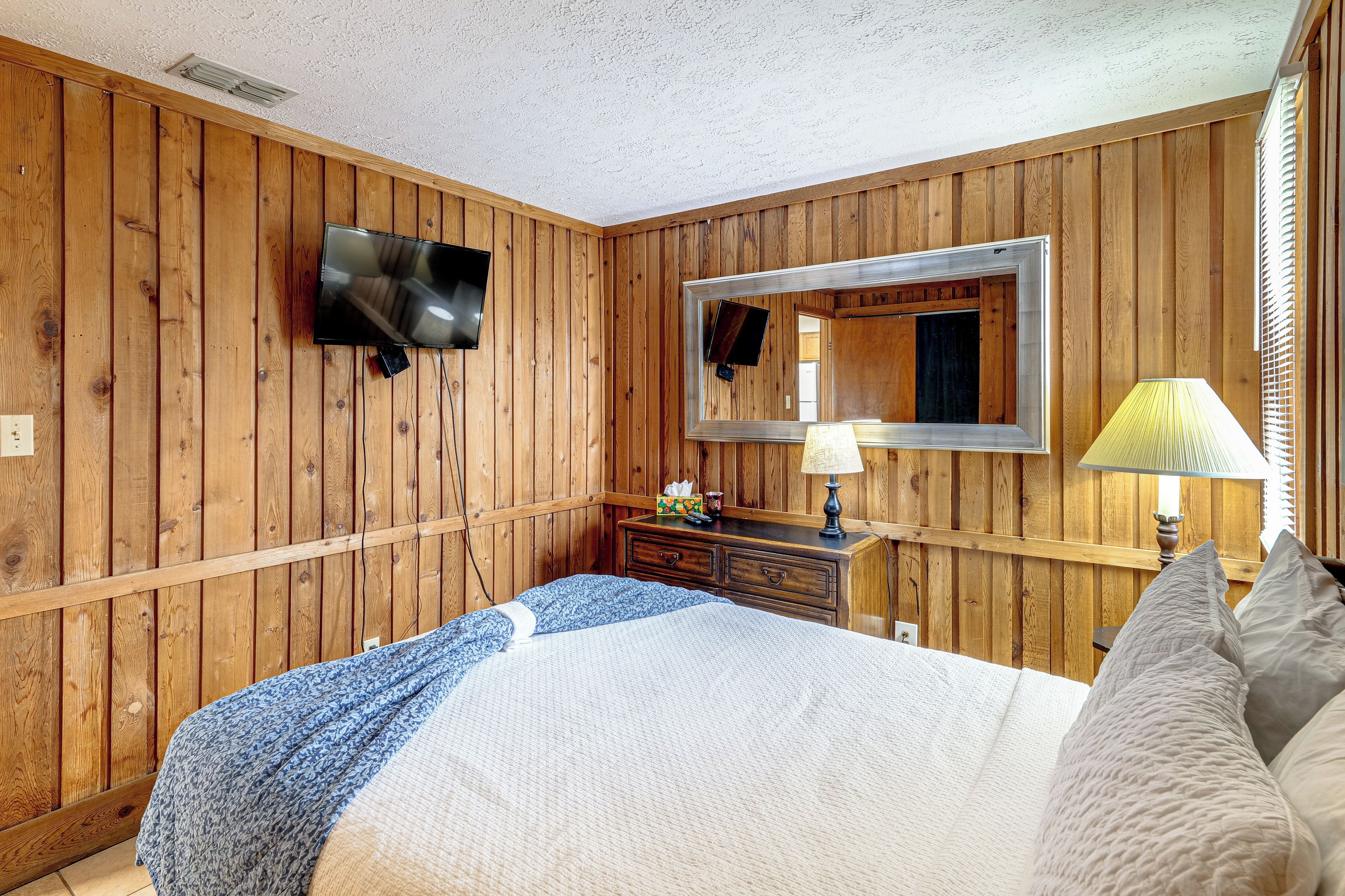 A room with wooden panel walls, a flat-screen TV mounted on the wall, a bedside table with a lamp, and a bed with a patterned blanket.