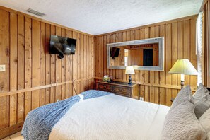 Ein Zimmer mit Holzwandverkleidung, einem Wand-Fernseher, einem Nachttisch mit Lampe und einem Bett mit gemustertem Bettbezug.