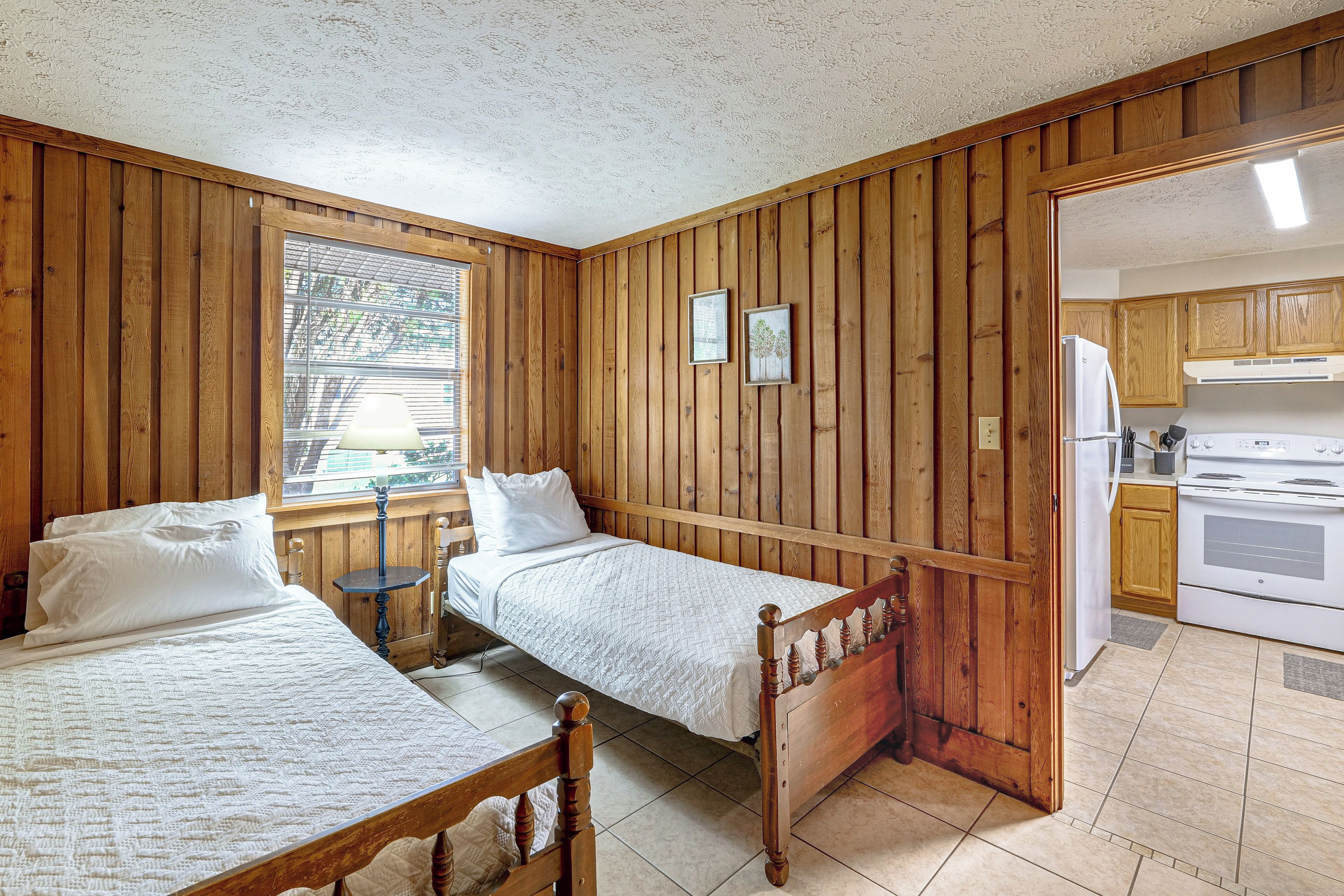 A room with two wooden beds, a window with blinds, a kitchen area with a stove, and wooden walls.