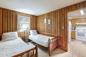 Ein Zimmer mit zwei Holzbetten, einem Fenster mit Jalousien, einer Küchenzeile mit Herd und Holzwänden.