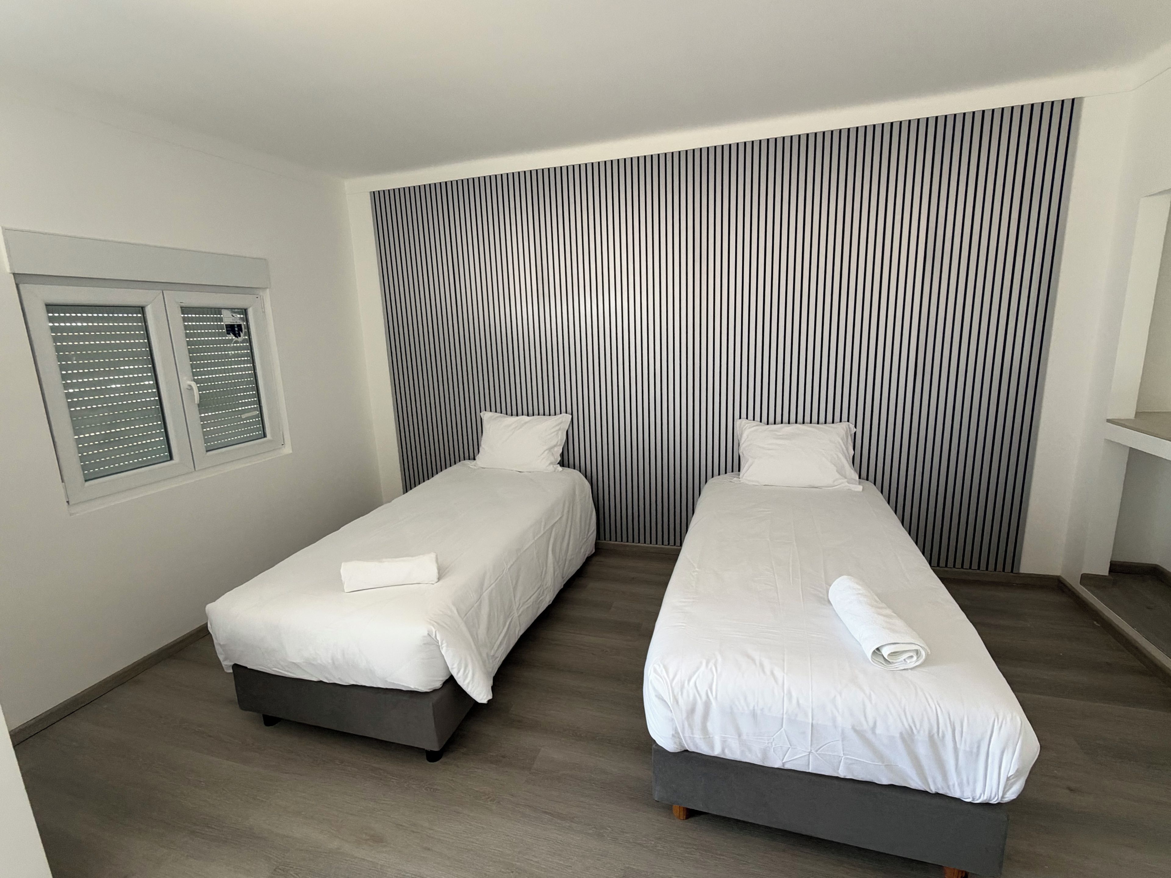 A hotel room with two single beds, a window with blinds, and a striped wall.