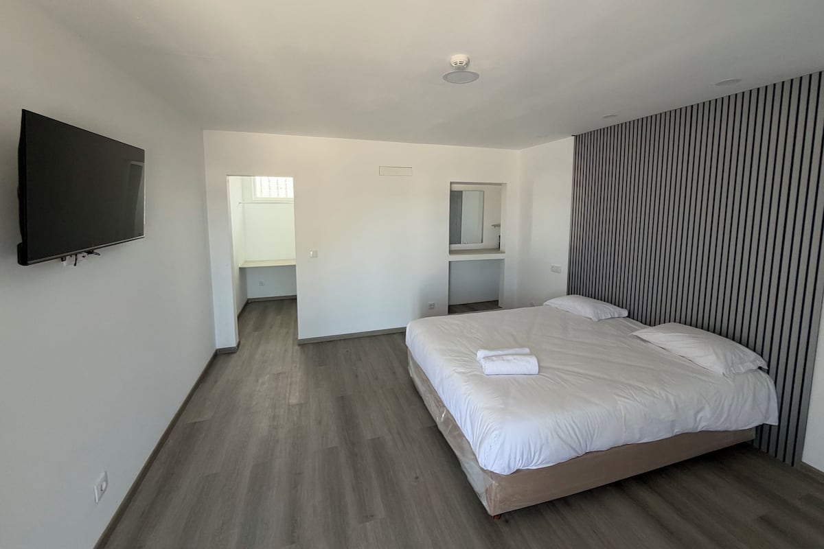 A hotel room with a large bed, a flat-screen TV mounted on the wall, a mirror above the bed, and a striped wall panel.