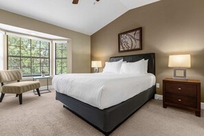 Quarto com cama grande, uma cadeira, mesa de cabeceira e janela com persianas.