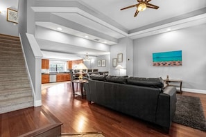 A spacious living area with a dark-colored sofa, a wooden staircase, a kitchen in the background, and a ceiling fan. - Family Getaway Near Disney With Pool and Games (Orlando)