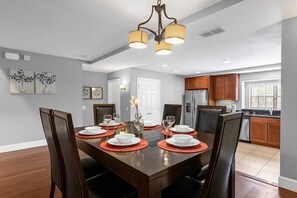 Uma sala de jantar com uma mesa de madeira posta para quatro pessoas, cercada por cadeiras. É possível ver uma cozinha ao fundo, com armários de madeira e uma geladeira.