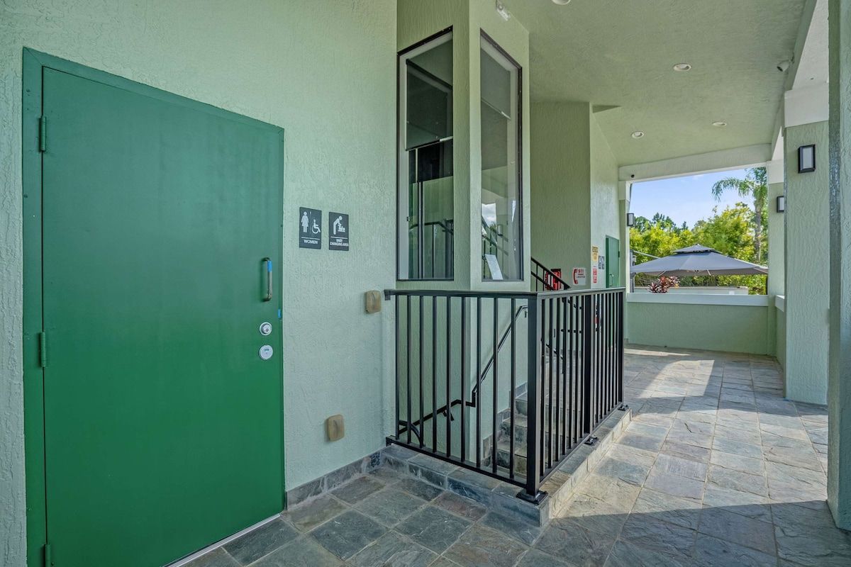 Un espacio al aire libre techado con una puerta verde, baranda metálica y vista a un estacionamiento.