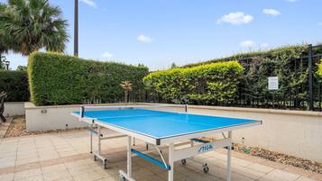 An outdoor ping pong table with a TIGA brand label, surrounded by a hedge and a palm tree.