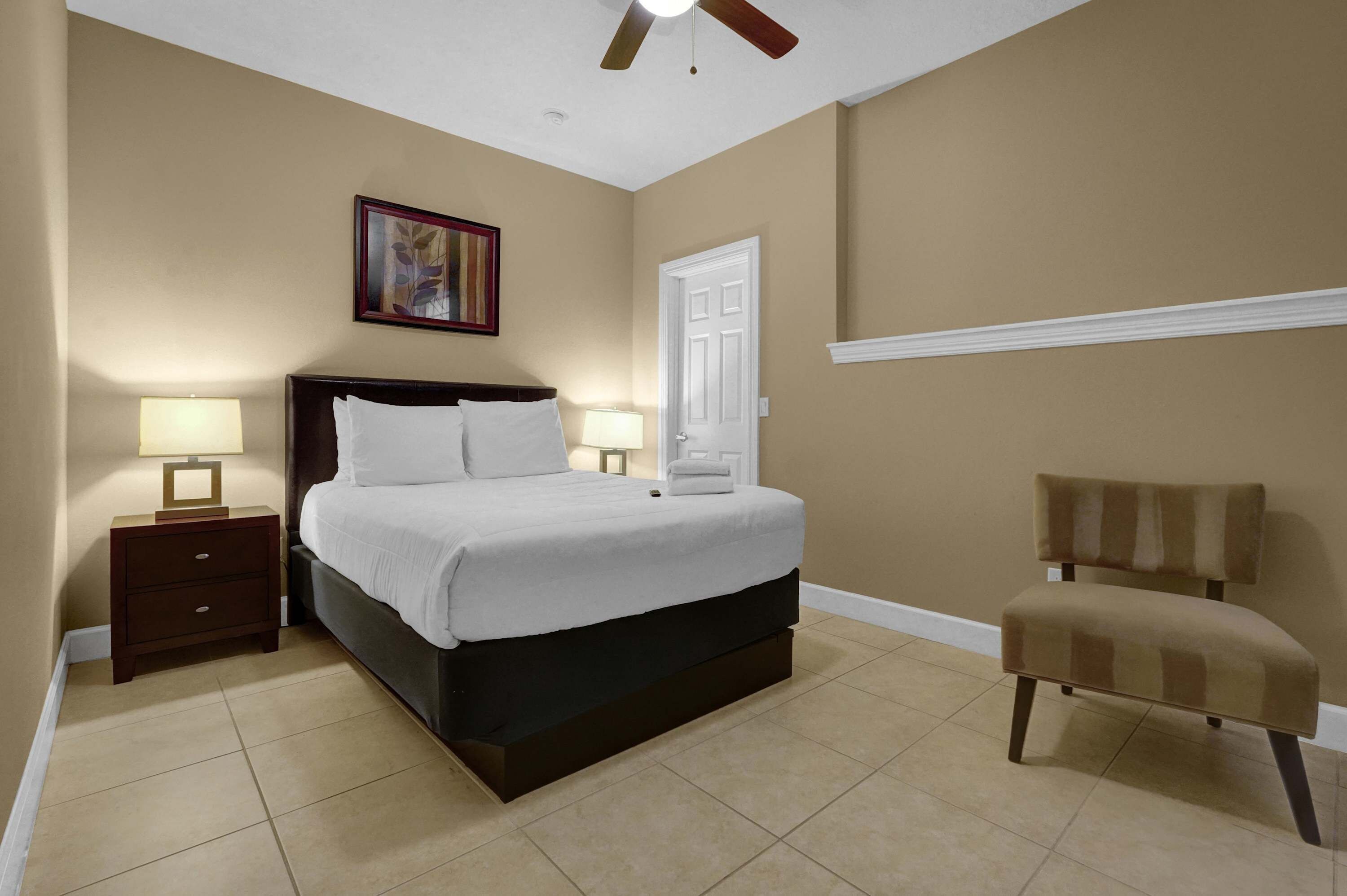 A bedroom with a bed, nightstands, a chair, a ceiling fan, a door, and a framed picture on the wall.