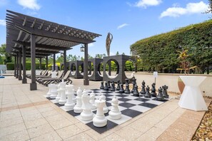 Un échiquier avec de grandes pièces disposées à l’extérieur, entouré de fauteuils et surmonté d’une pergola.