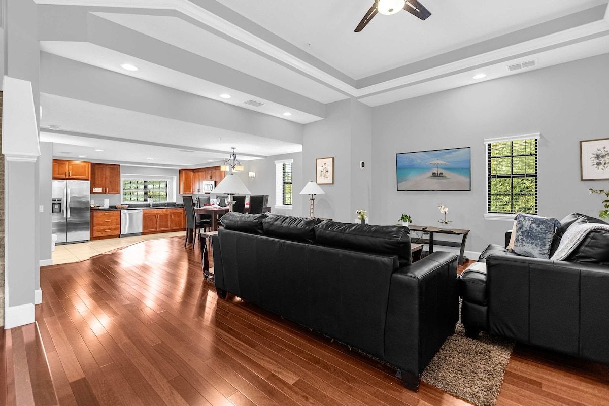Uma sala de estar ampla, com sofá de couro preto, piso de madeira e cozinha ao fundo.