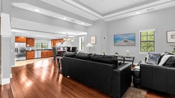 Uma sala de estar ampla, com sofá de couro preto, piso de madeira e cozinha ao fundo.
