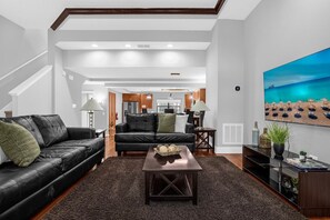 A modern living room with a black leather sofa, a dark coffee table, and a large painting of a beach scene. - Home Near Disney With Resort Pool Spa Access (Orlando)