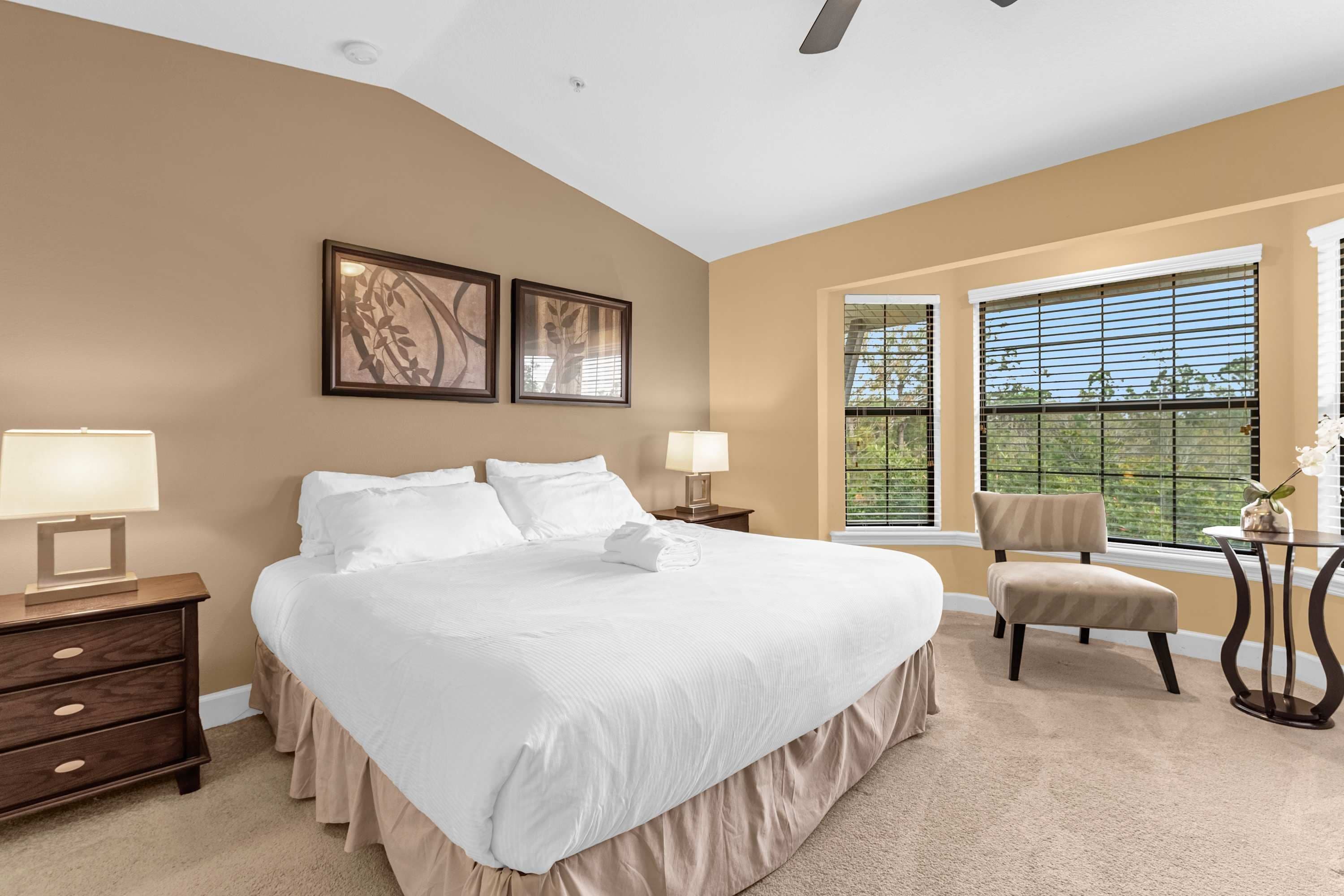 A bedroom with a large bed, two bedside lamps, a chair, a window with blinds, and artwork on the wall.