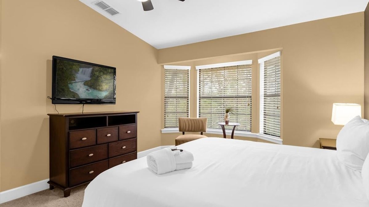 A hotel room with a bed, a television mounted on the wall, a dresser, a chair, a small table with a lamp, and a window with blinds.