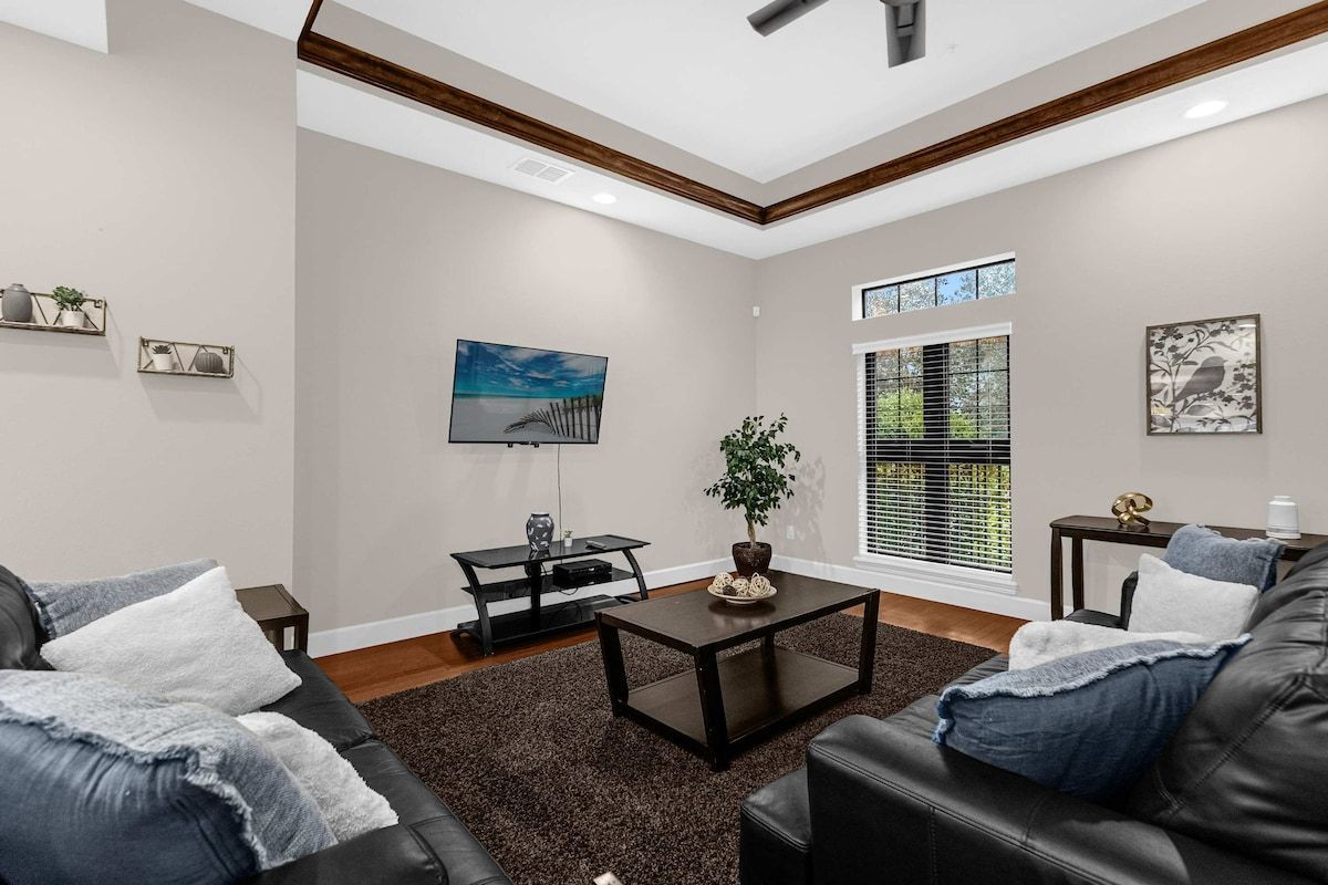 A living room with a sofa, coffee table, and TV. There is a window with blinds, a plant, and artwork on the walls.