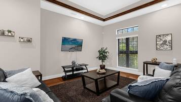 A living room with a sofa, coffee table, and TV. There is a window with blinds, a plant, and artwork on the walls.