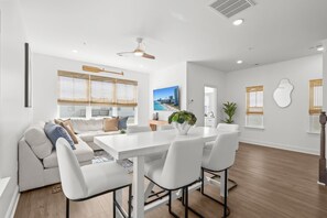 A modern living room with a white sofa, a dining table with chairs, a ceiling fan, and a large window with blinds. - Casa Bella | Family Coastal Retreat | Ocean Views (Myrtle Beach)