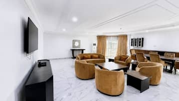 Una habitación de hotel moderna con televisión de pantalla plana, un conjunto de sofás, una mesa de comedor y un amplio ventanal con cortinas.