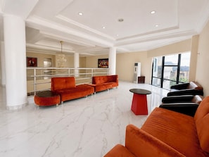 A spacious lobby with a marble floor, a red and black sofa set, a chandelier, and a large window offering a city view.