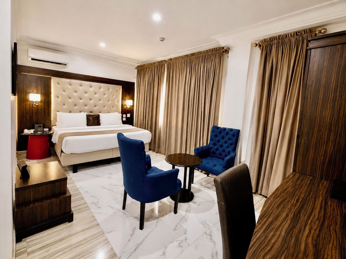 Une chambre d’hôtel avec un lit, deux fauteuils bleus, une petite table, un tabouret rouge et une table d’appoint en bois.