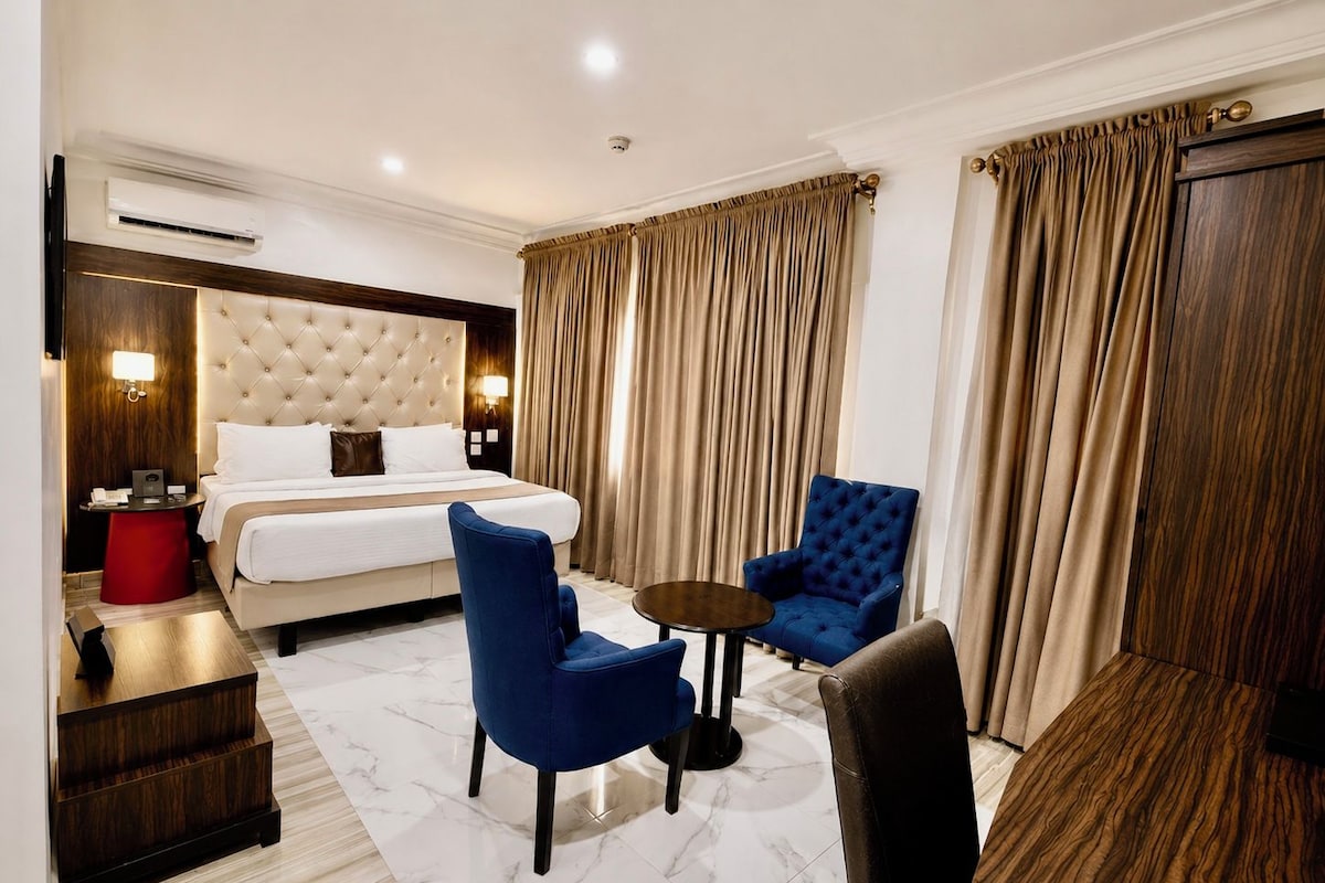 Een hotelkamer met een bed, twee blauwe fauteuils, een tafeltje, een rode kruk en een houten bijzettafeltje.