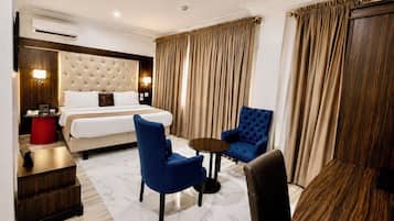 Une chambre d’hôtel avec un lit, deux fauteuils bleus, une petite table, un tabouret rouge et une table d’appoint en bois.