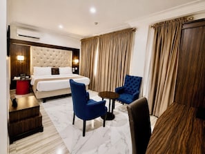 Een hotelkamer met een bed, twee blauwe fauteuils, een tafeltje, een rode kruk en een houten bijzettafeltje.