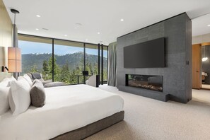 A modern bedroom with a large bed, a fireplace, a TV mounted on the wall, and a view of mountains through large windows. - Just Listed: New Ski Residence, Views, Gym, Hot tub (Whistler)