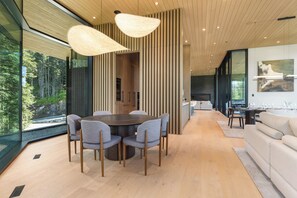 A modern interior with a dining area, a sofa, and a large window offering a forest view. - Just Listed: New Ski Residence, Views, Gym, Hot tub (Whistler)