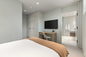 A modern bedroom with a bed, a desk, a flat-screen TV, and a bathroom visible through an open door. - Just Listed: New Ski Residence, Views, Gym, Hot tub (Whistler)