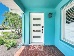 A turquoise house with a white door, a small porch with a red sun-patterned mat, and a black wall-mounted light. - Luxury Florida Retreat with Pool & Exceptional Outdoor Living (St. Petersburg)